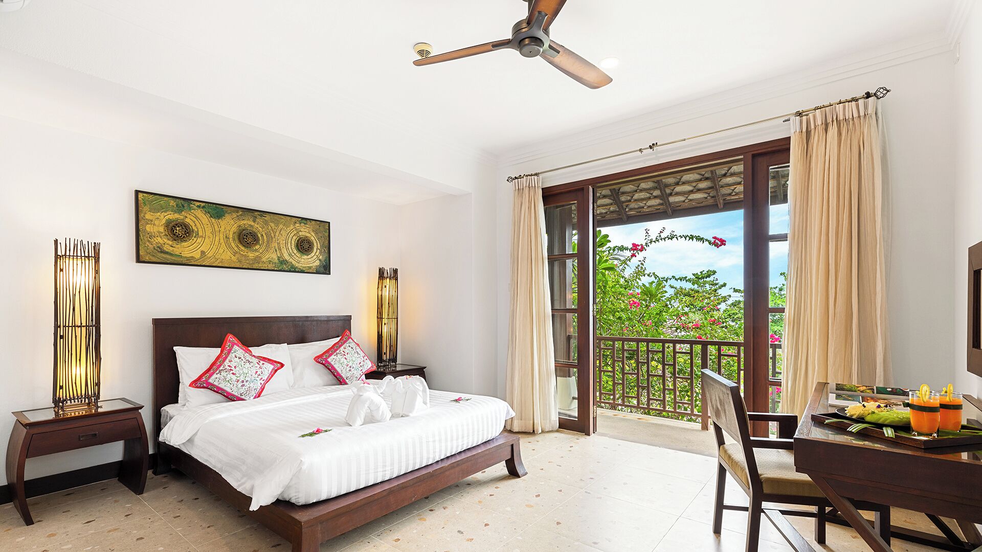 Une chambre avec un lit, un ventilateur de plafond, une table à manger avec une chaise, et un balcon donnant sur un paysage verdoyant.