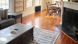 A spacious living room with a fireplace, wooden flooring, and a dining area with chairs and a table. - Quissett Landing | Sleeps 12 | Walk to The Knob (Falmouth)