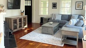 A living room with a grey sofa, a coffee table, a TV mounted on the wall, and a wooden cabinet. - Quissett Landing | Sleeps 12 | Walk to The Knob (Falmouth)