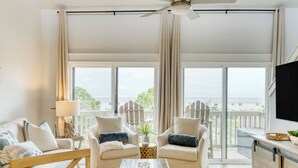 A spacious living room with a sofa, two armchairs, a coffee table, and a TV. Large windows offer a view of the outdoors.