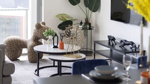 A modern living room with a round dining table, a large potted plant, a clock on the wall, and a TV mounted on the wall.