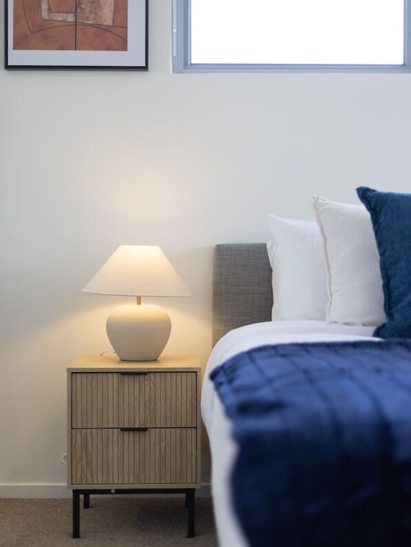 A neatly arranged bedroom with a bed, a bedside table with a lamp, and a framed picture on the wall.
