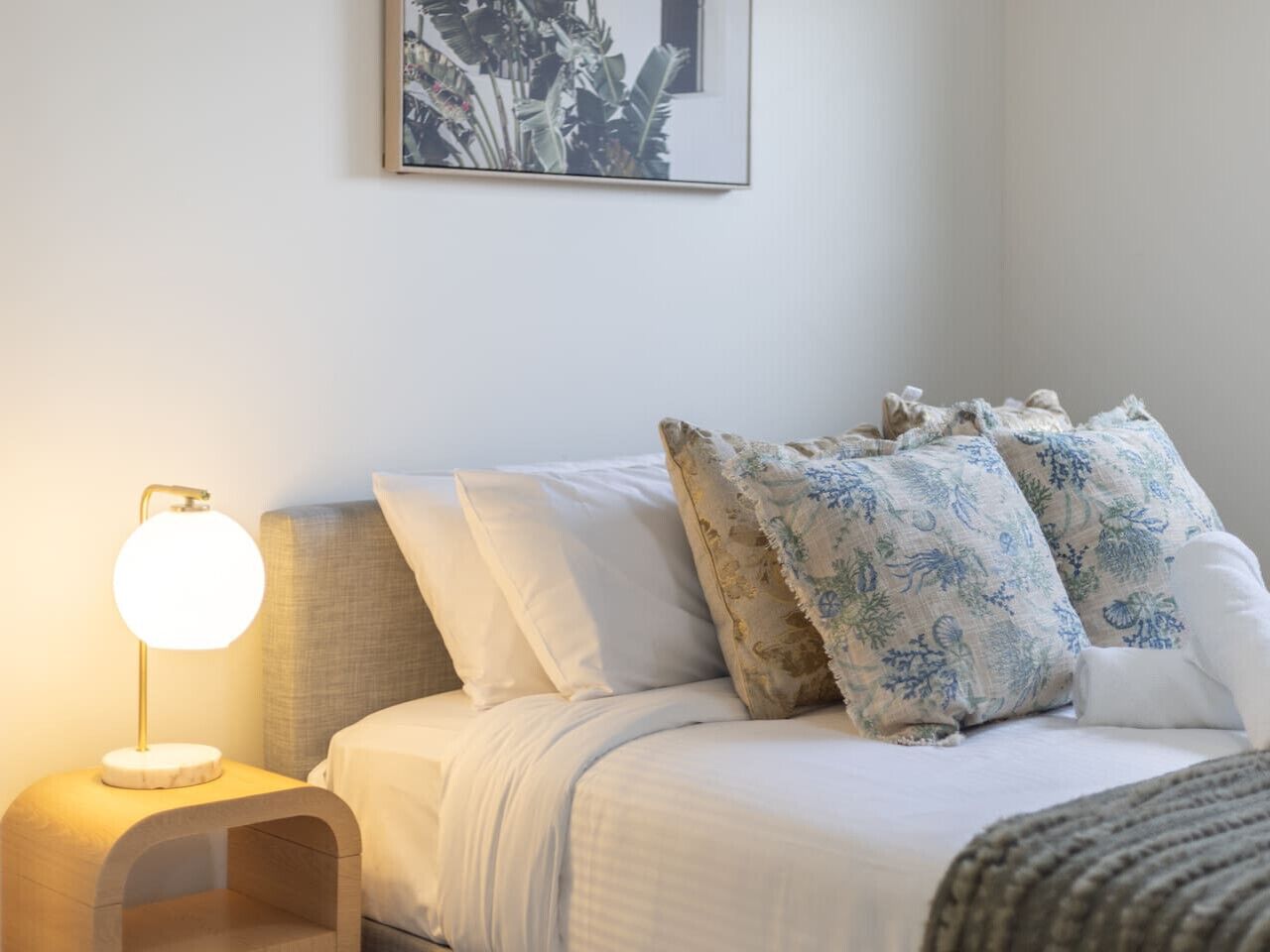 A neatly made bed with white bedding and blue floral pillows, a bedside lamp, and a framed picture on the wall.