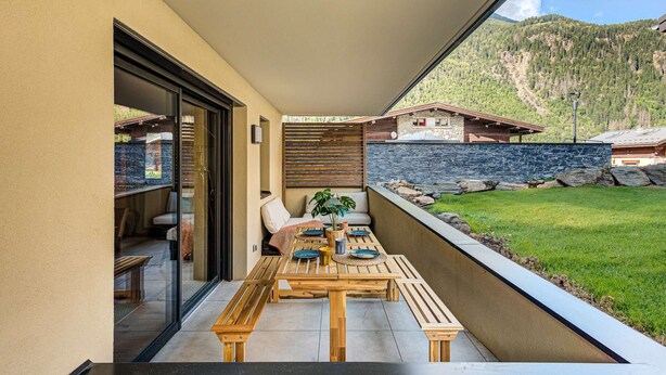 Un espacio moderno para comer al aire libre con mesa y bancos de madera, puerta de vidrio que da a un porche cubierto, y vistas a un paisaje montañoso.