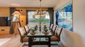 Comedor con mesa de madera, seis sillas y una araña. Se observa una ventana grande con vista a un edificio y paisaje.