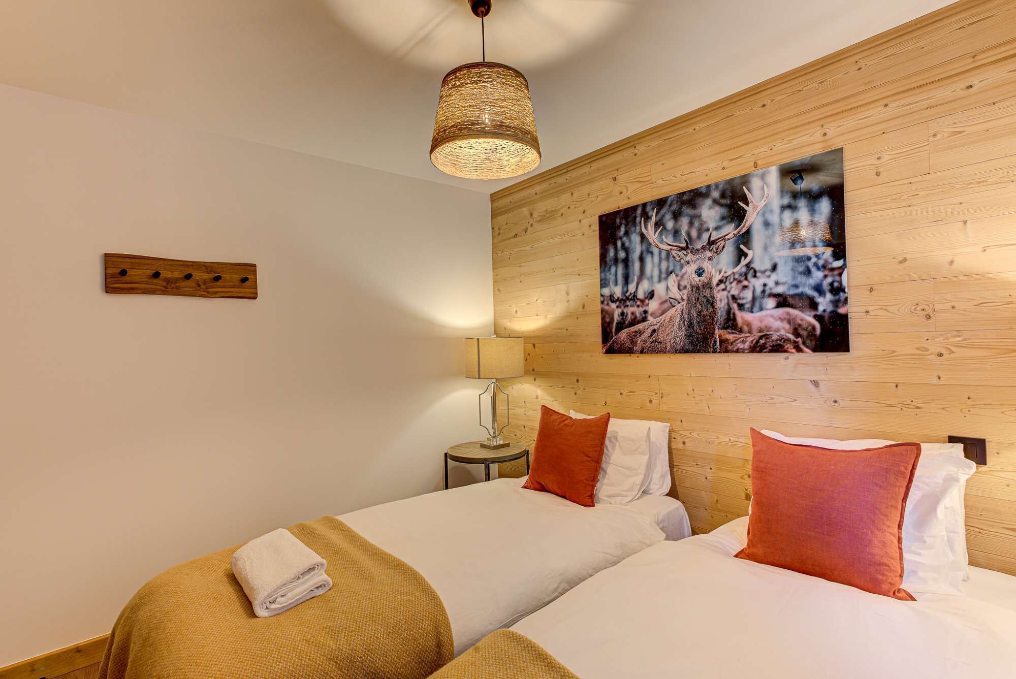 Habitación de hotel con dos camas, cabecero de madera, un cuadro colgado en la pared con ciervos, lámpara colgante y una mesita con lámpara.