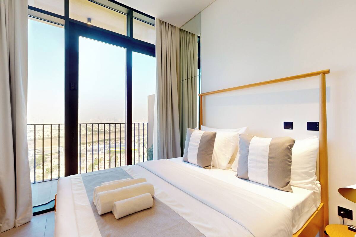 Une chambre d’hôtel moderne dotée d’un grand lit, offrant une vue sur la ville grâce à de larges fenêtres du sol au plafond, et disposant d’un balcon avec une balustrade.
