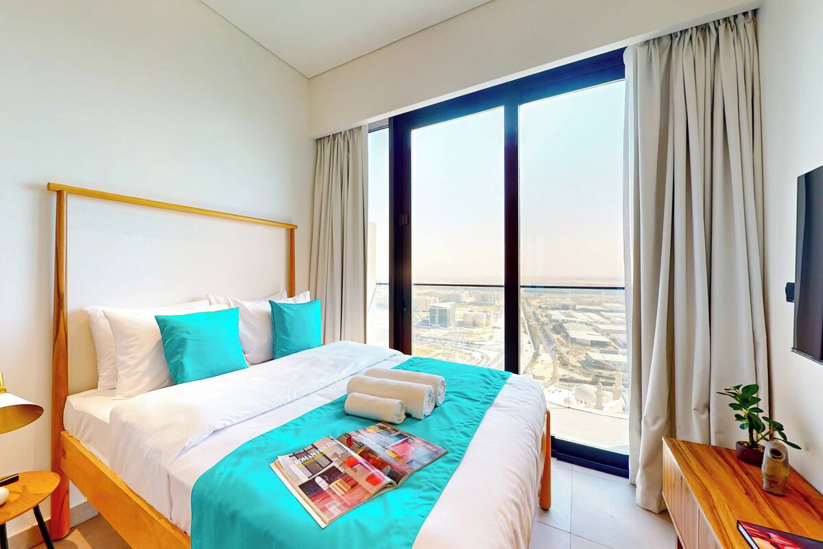 Une chambre d’hôtel moderne avec un grand lit, une table de chevet, une lampe, un magazine et une vue sur la ville grâce à de grandes fenêtres.