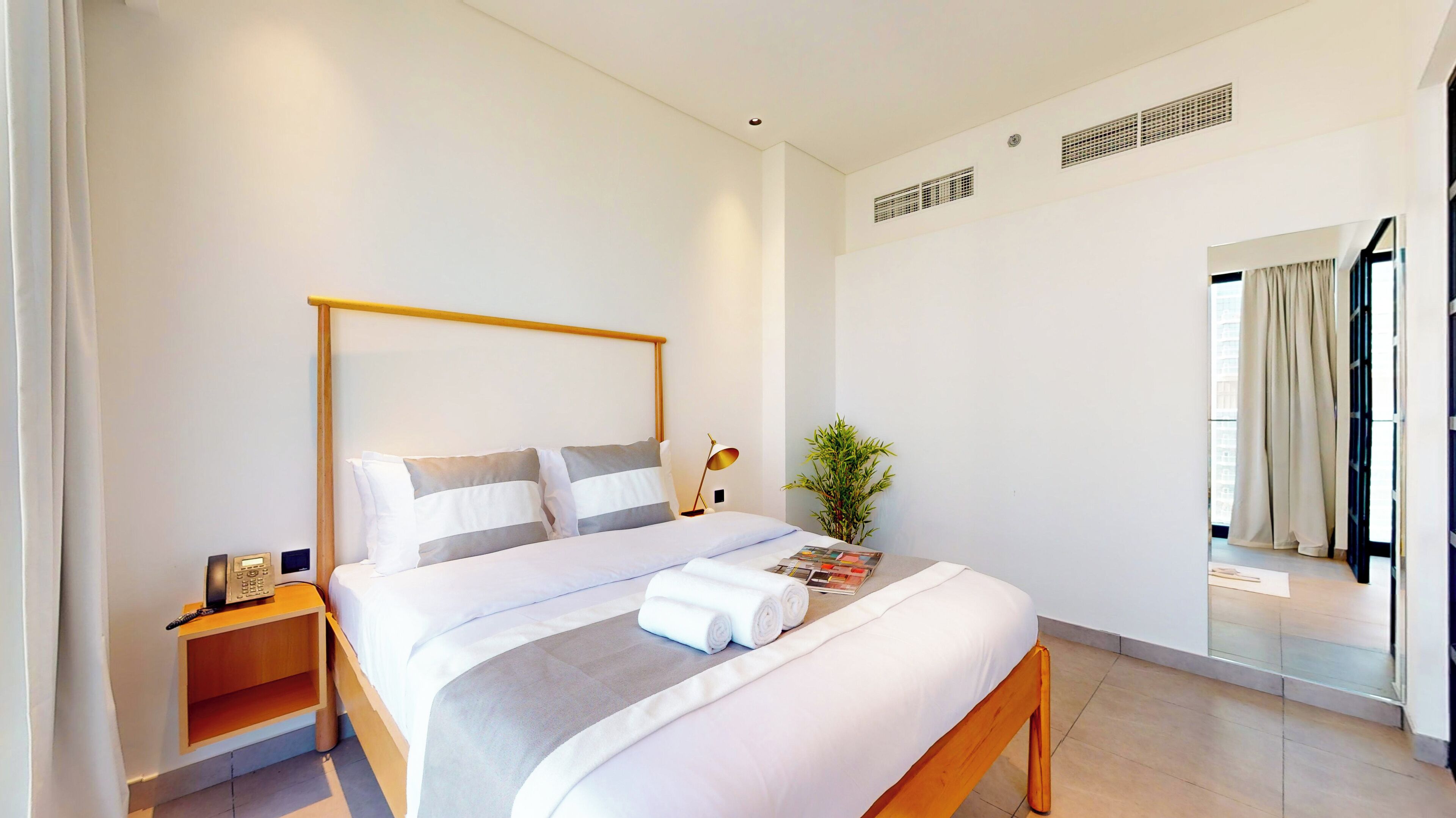 Una habitación de hotel moderna con estructura de cama de madera, ropa de cama blanca, una mesita de noche con lámpara y una pequeña planta.