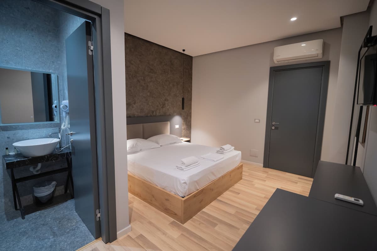 A modern bedroom with a wooden bed, a built-in nightstand, a bedside table, a large mirror, and a bathroom with a sink and towel rack.