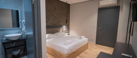 A modern bedroom with a wooden bed, a built-in nightstand, a bedside table, a large mirror, and a bathroom with a sink and towel rack.