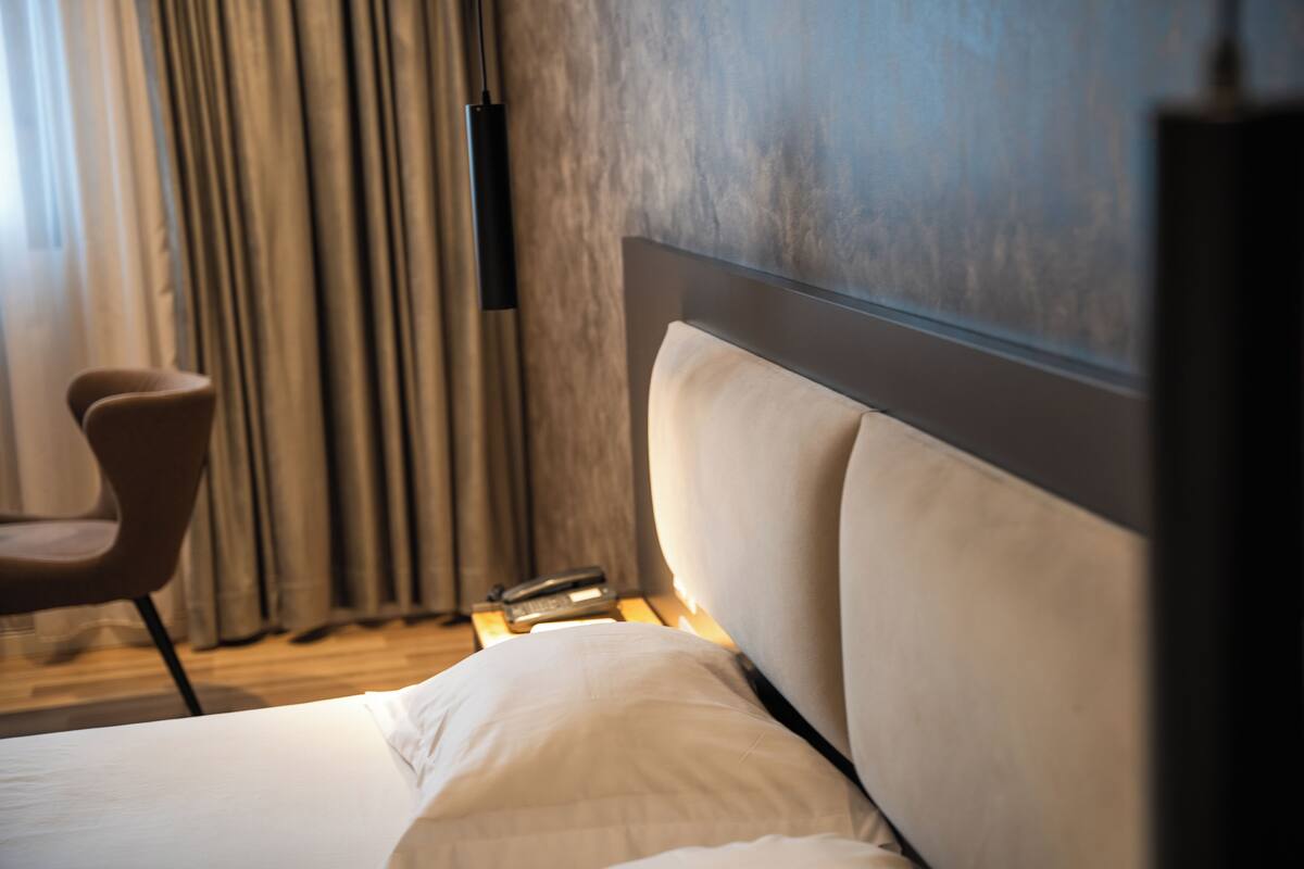 A hotel room with a bed, a chair, a bedside table, and a wall-mounted phone.