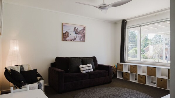 A living room with a dark brown sofa, a white table, and a lamp. There is a window with a view of houses and trees outside.