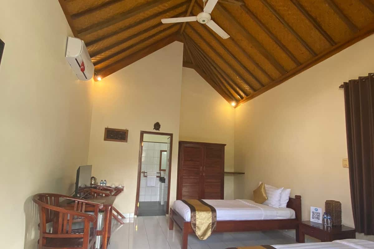 A hotel room with a wooden ceiling, a bed, a desk with chairs, and a bathroom visible through an open door.