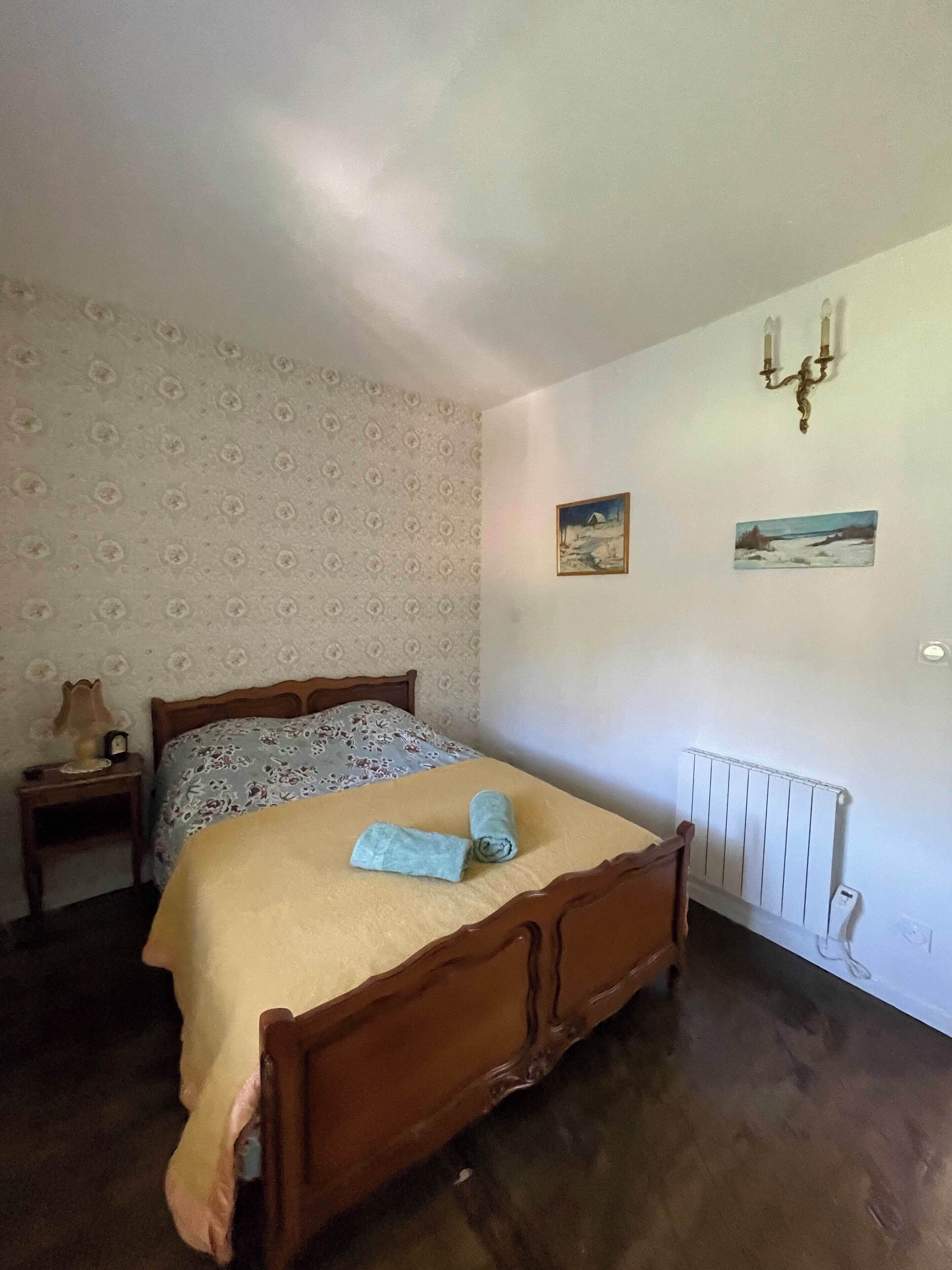 A bedroom with a wooden bed, a nightstand with a lamp, patterned wallpaper, and two wall-mounted pictures.