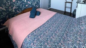 A bedroom with floral wallpaper, a bed with a pink and blue floral bedspread, a wooden headboard, a white door, a small table with a lamp, and a picture on the wall.