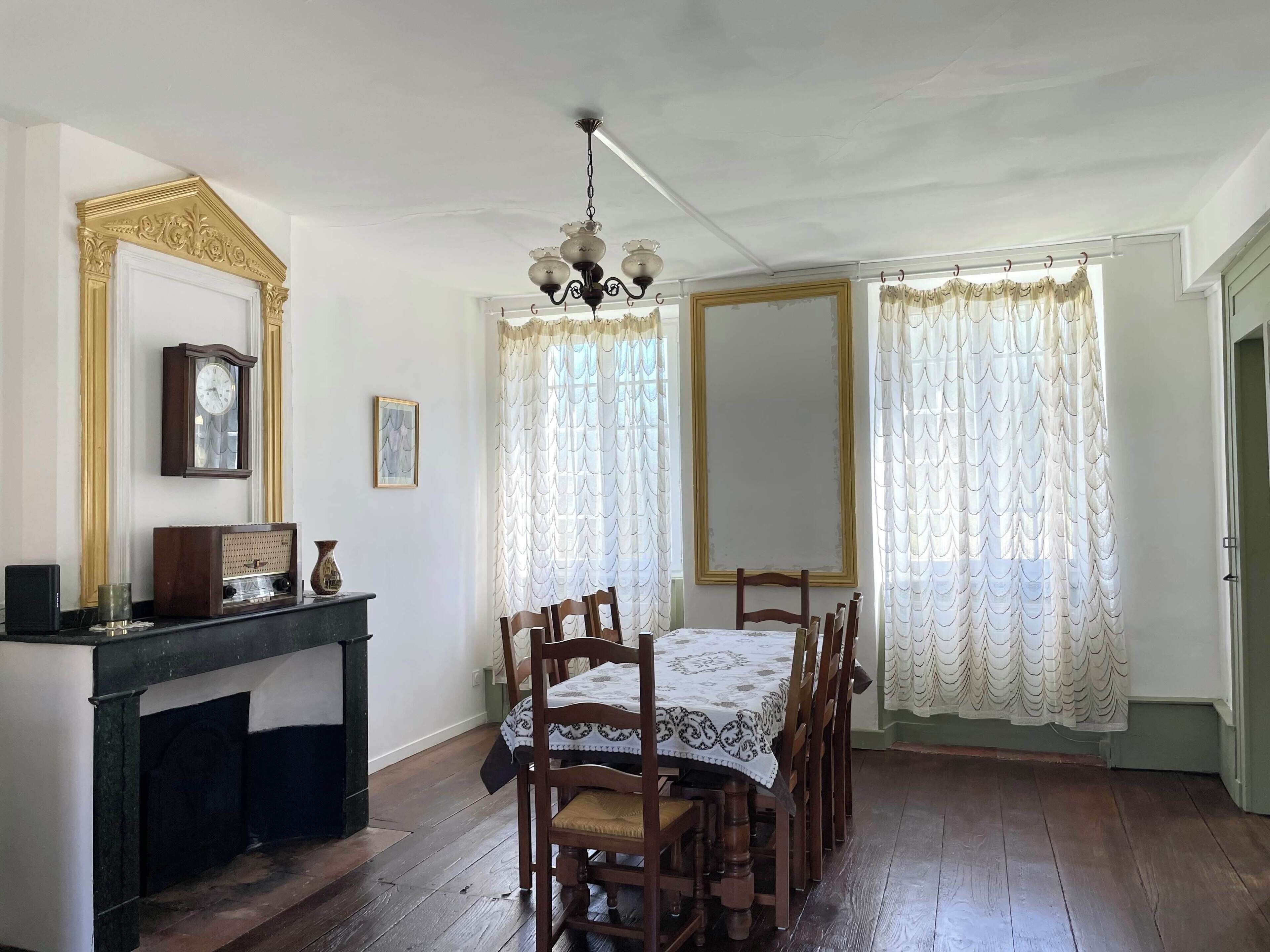 Een eetkamer met een houten tafel en stoelen, een open haard, een klok en een spiegel aan de muur.