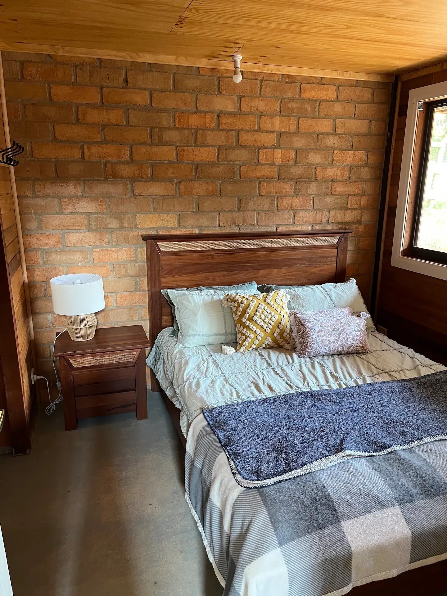 Une chambre avec un mur en briques, une tête de lit en bois, une table de chevet avec une lampe et une fenêtre.