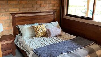 A bedroom with a wooden headboard, a brick wall, a window with blinds, and a bed with multiple pillows and a blanket.