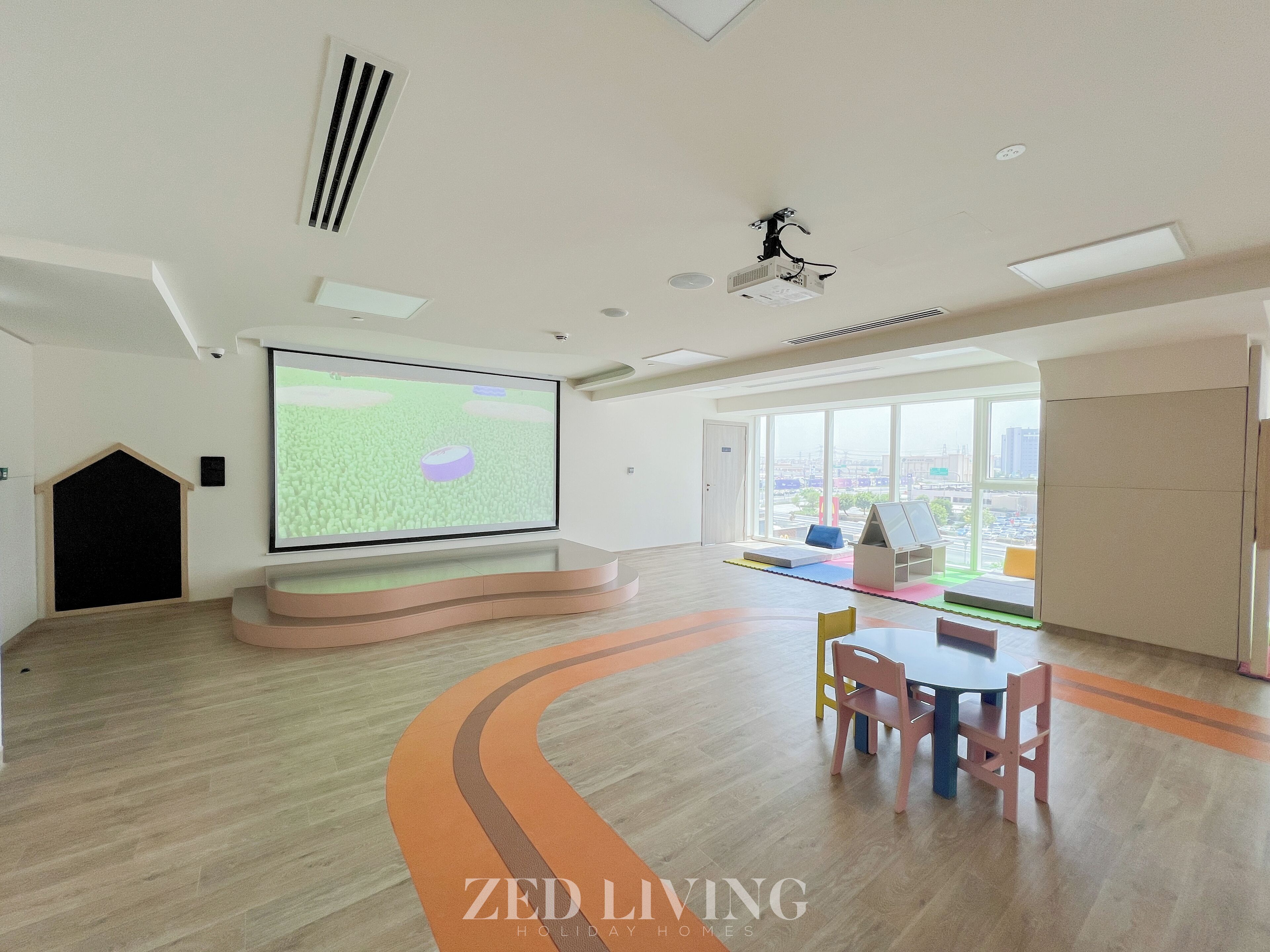 Una habitación amplia con una pantalla grande para presentaciones, un área de juegos con colchonetas coloridas y vistas a la ciudad a través de amplios ventanales.