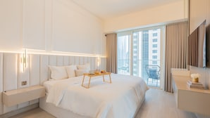Habitación de hotel moderna con una cama grande, mesitas de noche, lámparas de noche, una pequeña mesa con artículos de desayuno, una silla, un televisor y un amplio ventanal con cortinas.