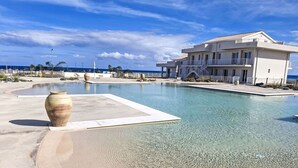 A resort pool with clear water, a sandy beach, and a large building with balconies. - Lovely apartment for 4 guests with WIFI, pool, A/C, TV and terrace (Pachino)