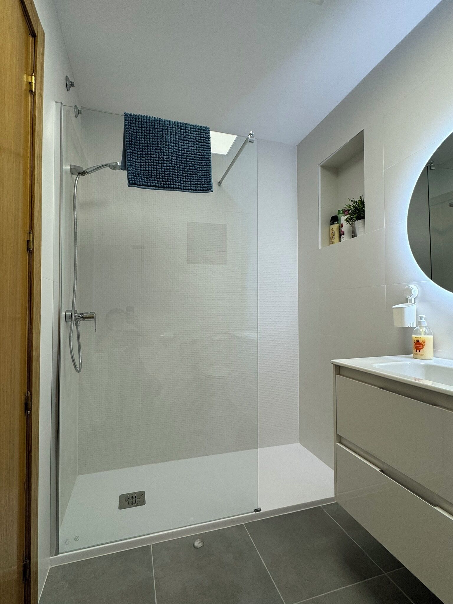 Une salle de bain moderne avec une douche à l’italienne en verre, un meuble-lavabo blanc avec un miroir rond et une petite étagère contenant des articles de toilette.