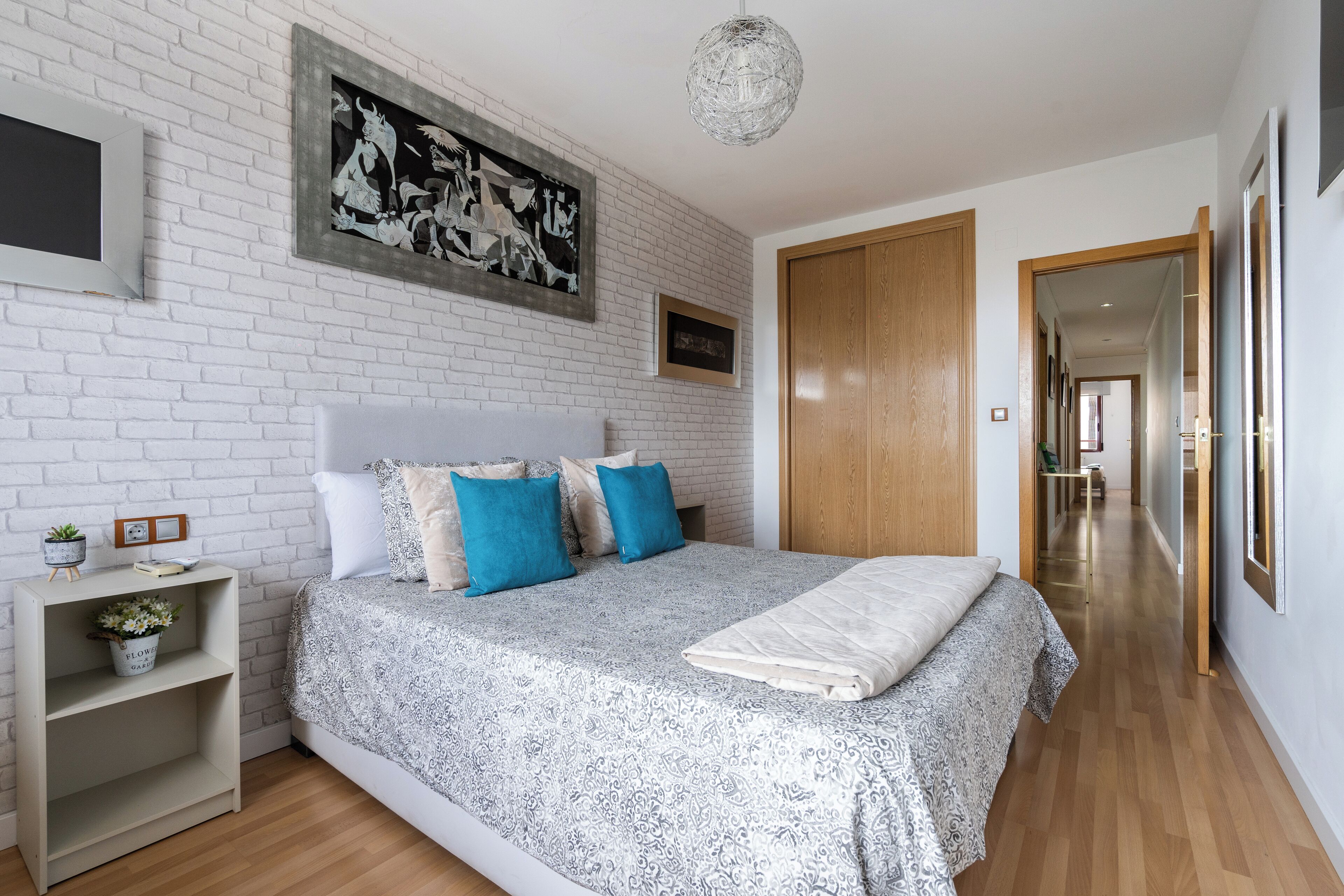 Un dormitorio moderno con pared de ladrillo blanco, una cama grande con ropa de cama estampada, una puerta de madera y un pasillo que conduce a otra habitación.
