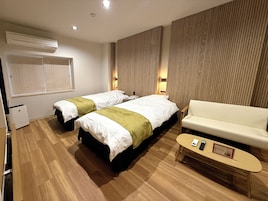 A hotel room with two beds, a sofa, a small table, and a wall-mounted air conditioner.