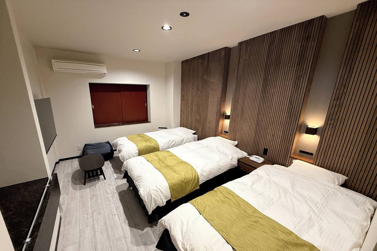 Ein Hotelzimmer mit zwei Betten, einem kleinen Tisch und einem Stuhl. Ein Fenster mit rotem Rollo, eine Wand-Klimaanlage und eine Wandverkleidung aus Holz sind zu sehen.