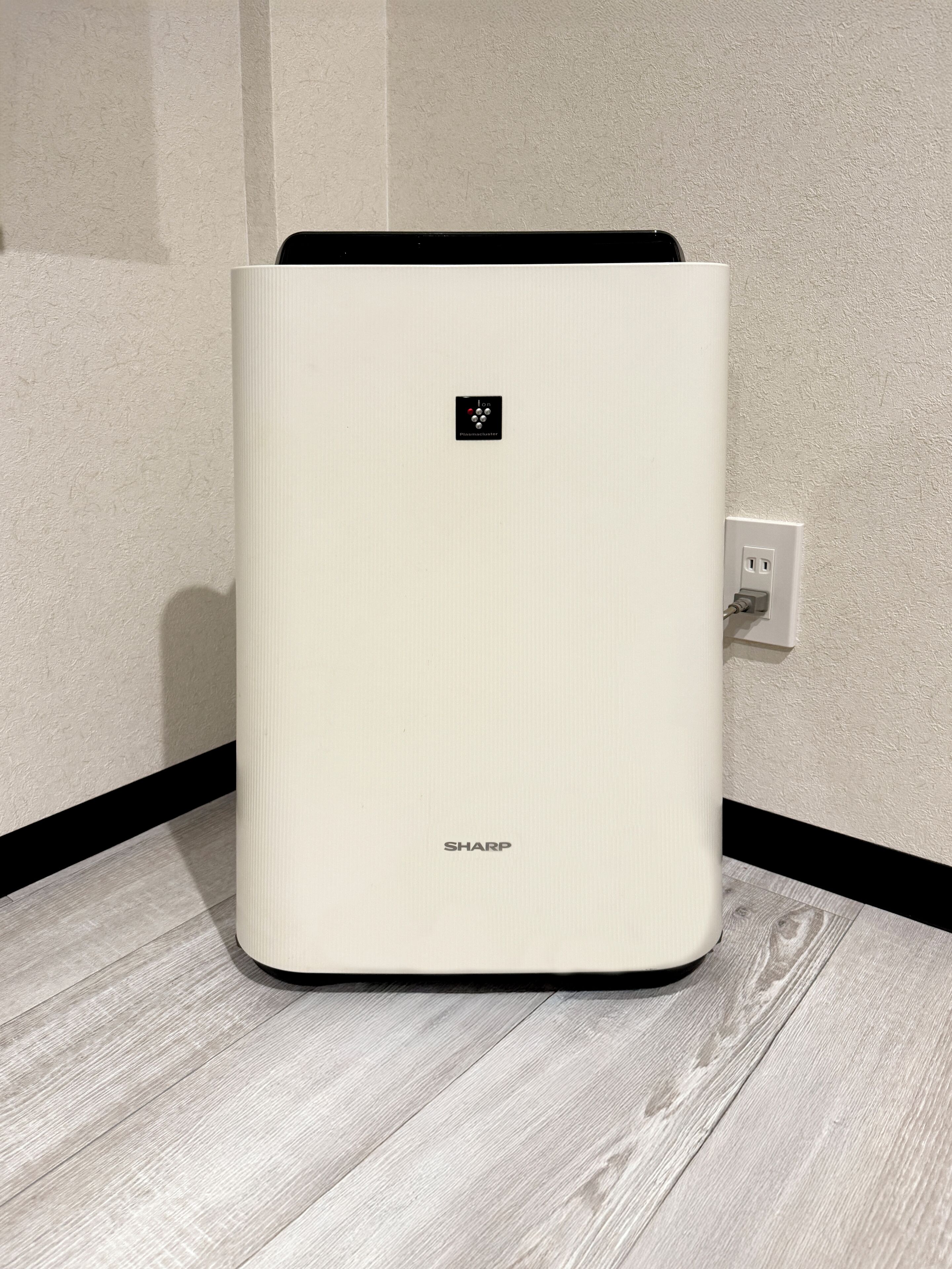 A white, modern, rectangular device with a black top, labeled "SHARP," placed on a wooden floor against a textured wall.