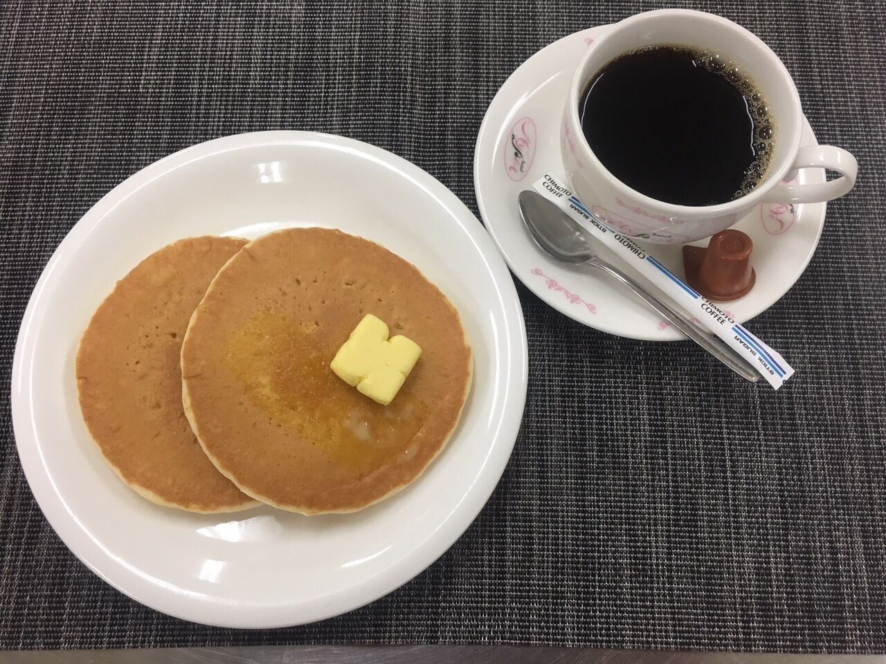Un plato con dos tortitas, un trozo de mantequilla, una taza de café y una cuchara sobre una mesa.