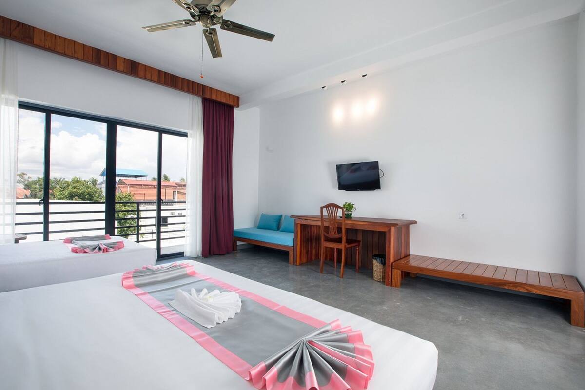 A hotel room with a large bed, a desk, a TV, and a balcony with a view of buildings.