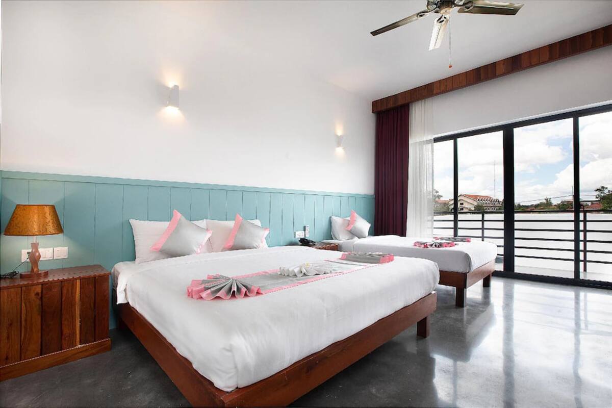 A hotel room with a large bed, a smaller bed, a nightstand, a lamp, and a view of the outside through large windows.