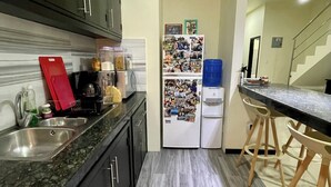 A kitchen with dark cabinets, a granite countertop, a refrigerator covered with photos, a sink, and a wooden stool.