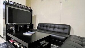 A living room with a black leather sofa, a black coffee table, a flat-screen TV on a stand, and an air conditioning unit mounted on the wall.