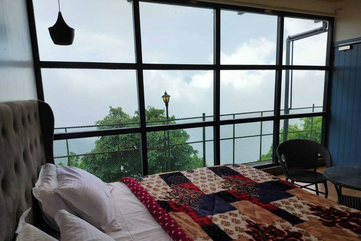 A bedroom with a large window, a bed with white bedding, a colorful quilt, and a chair by the window.