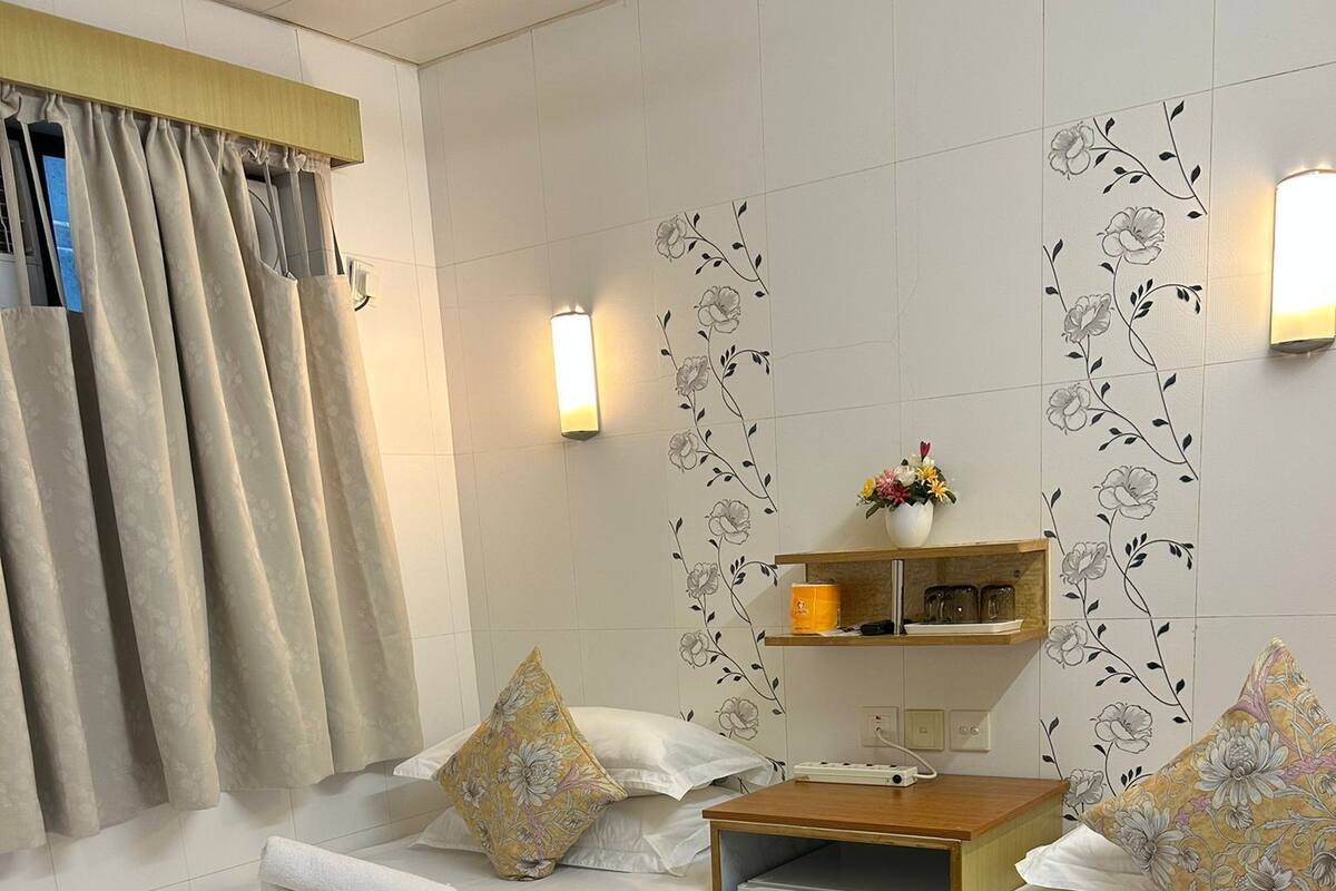 A hotel room with two beds, a small refrigerator, a wooden nightstand, a wall-mounted shelf with decorative items, and a window with curtains.