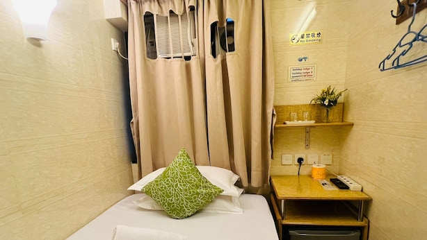 A small, neatly made bed with a green pillow and a rolled white towel, a small table with a phone and a cup, and a wall-mounted shelf with a plant. - HONG KONG SUN GUEST HOUSE (Kowloon)