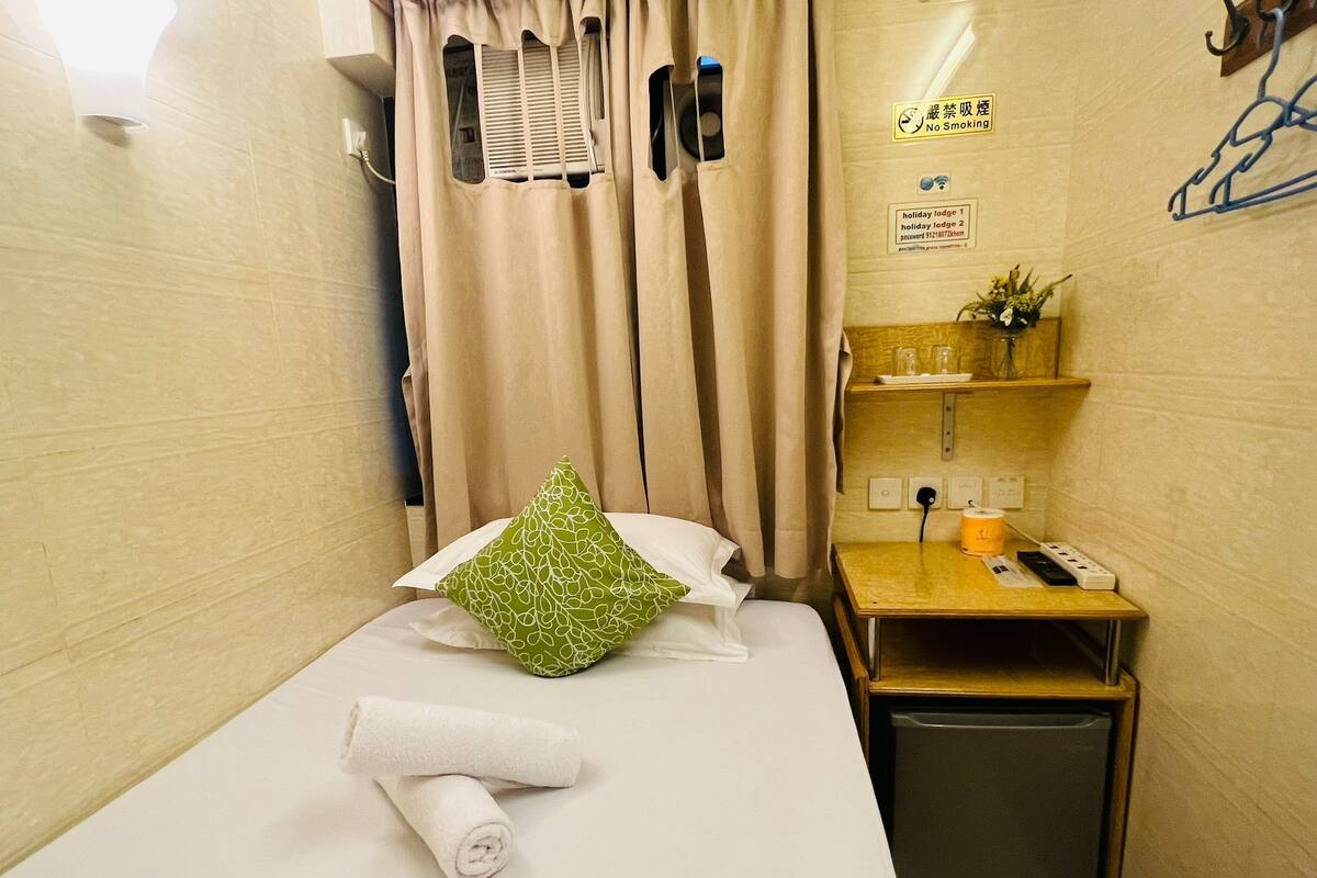 A small, neatly made bed with a green pillow and a rolled white towel, a small table with a phone and a cup, and a wall-mounted shelf with a plant.