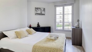 A neatly made bed with patterned pillows, a small statue on a nightstand, a picture of the Eiffel Tower, and a window with a view of buildings. - 123home -The Outlet (Serris)