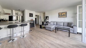 A modern apartment with a kitchen, dining area, and living room. It features a grey sofa, a coffee table, and a bookshelf. - 123home -The Outlet (Serris)