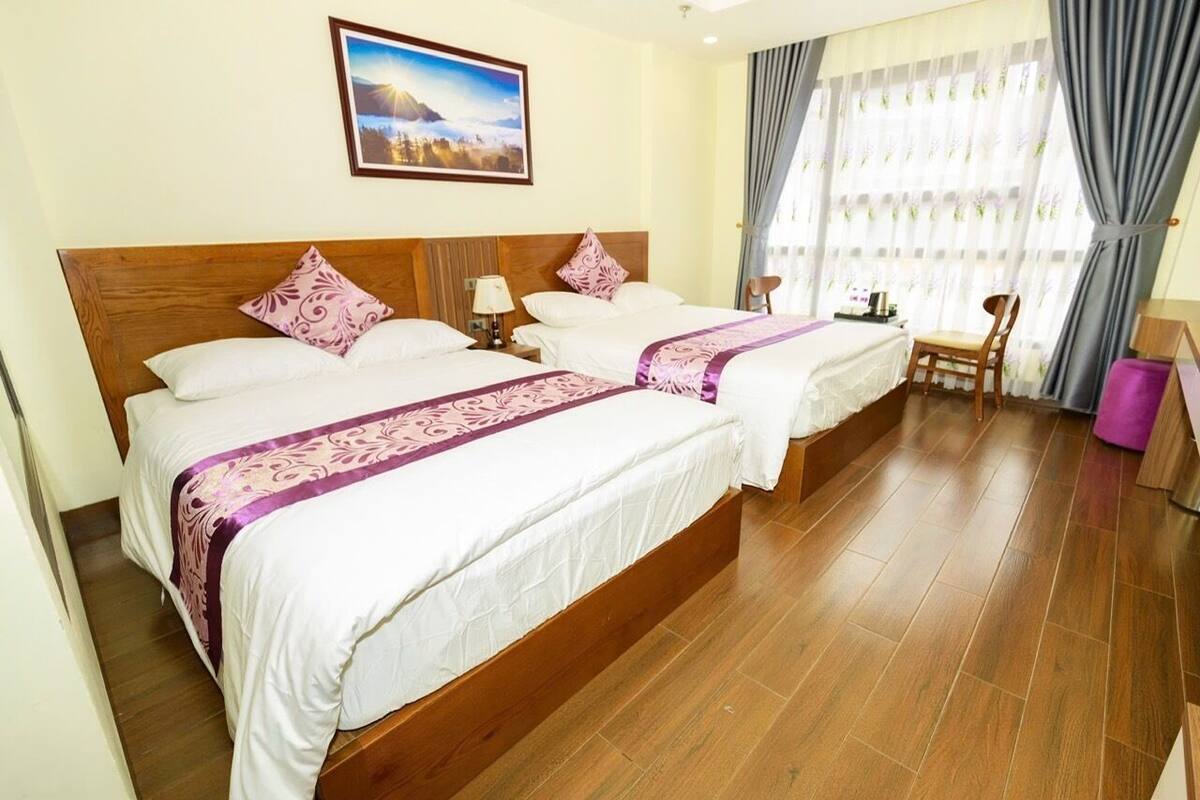 A hotel room with two beds, wooden headboards, a wooden floor, a chair, a table, and a window with curtains.