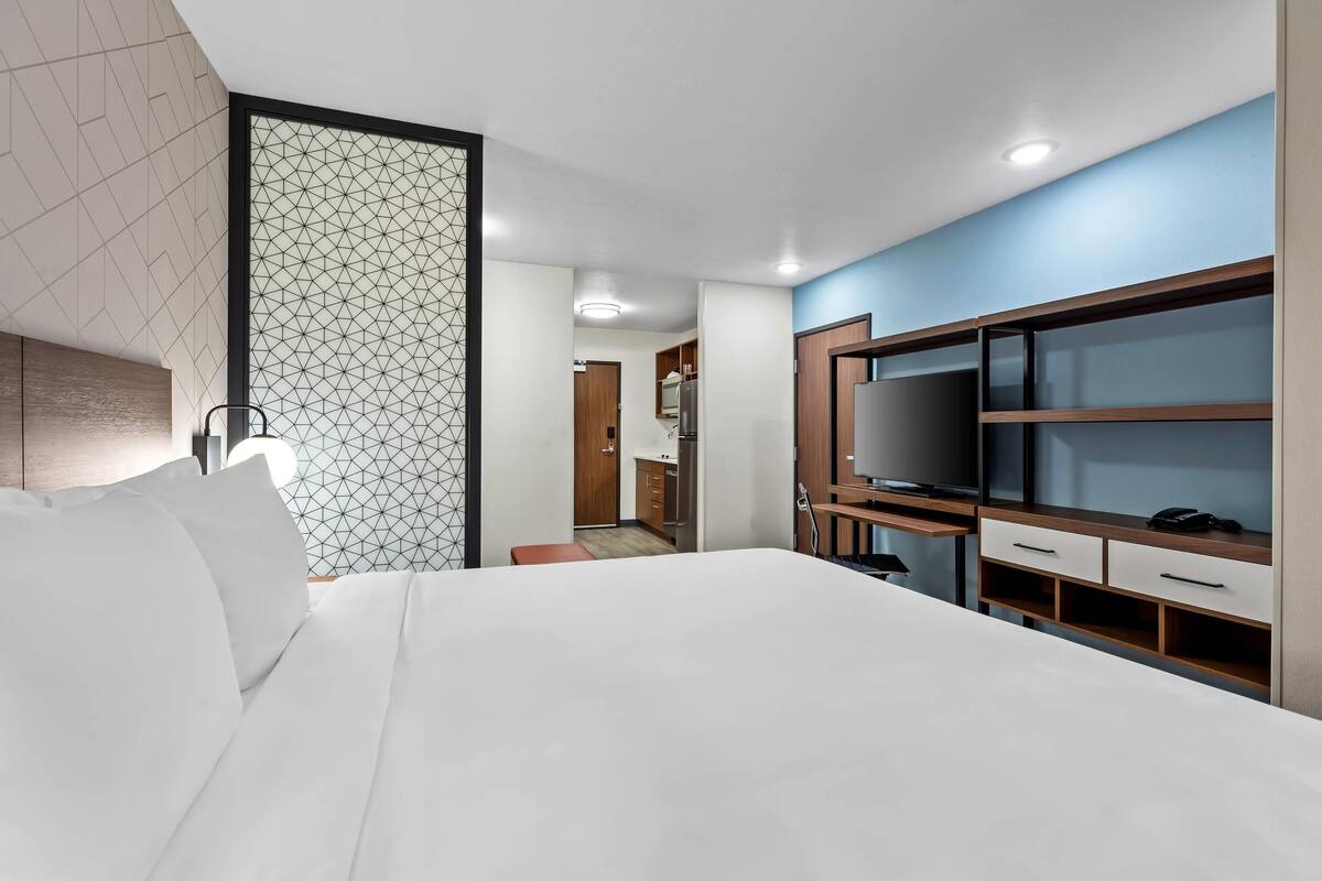 A modern hotel room with a large bed, a TV, a desk, and a kitchenette.