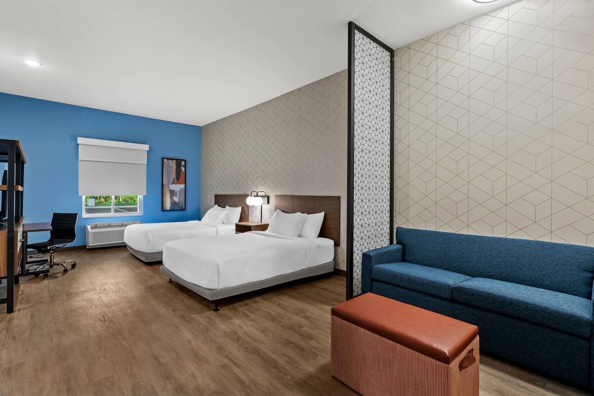 A hotel room with two beds, a blue sofa, a brown ottoman, a desk with a chair, and a window with blinds.
