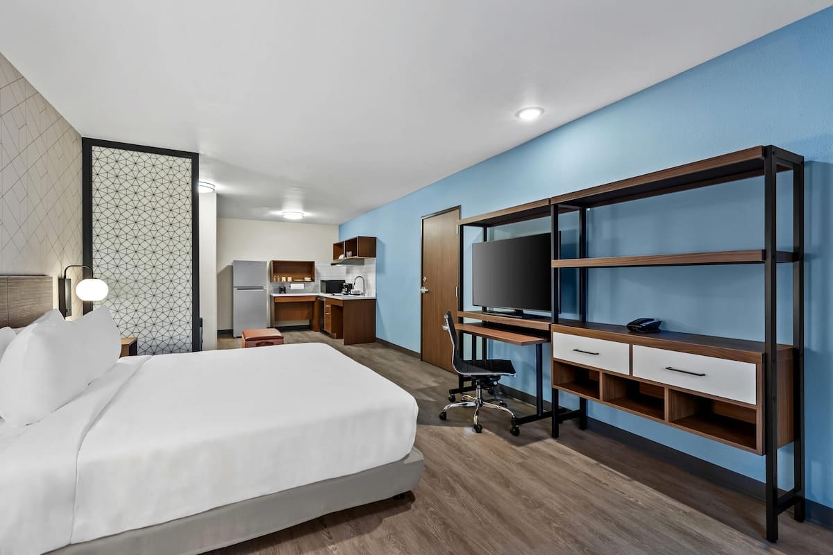 A hotel room with a bed, desk, TV, and kitchenette.