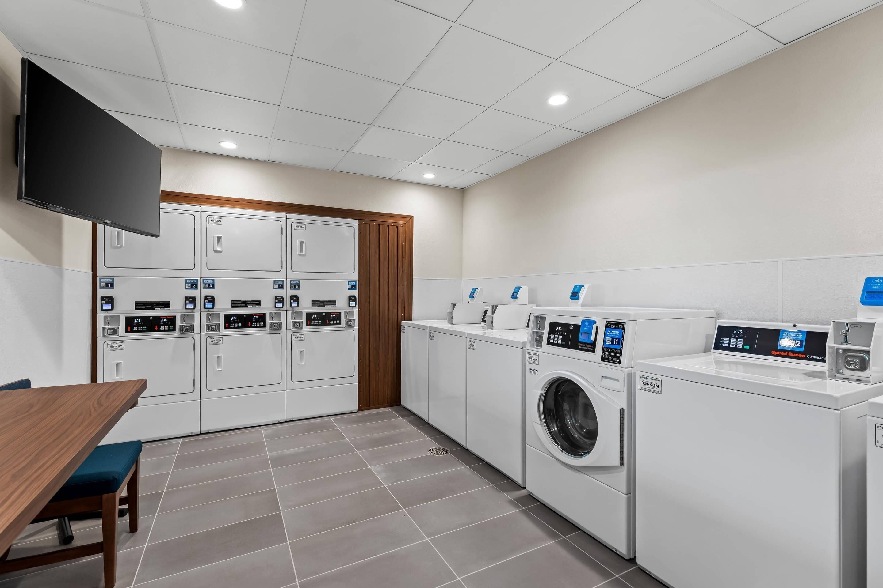 A laundry room with multiple washing machines and dryers, a wooden table, and a flat-screen TV mounted on the wall.