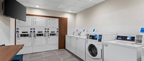 A laundry room with multiple washing machines and dryers, a wooden table, and a flat-screen TV mounted on the wall.