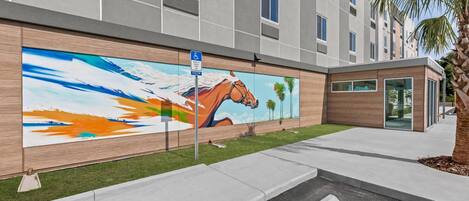 Un edificio moderno con un mural de un caballo y una pintura abstracta, un cartel de estacionamiento y una palmera.