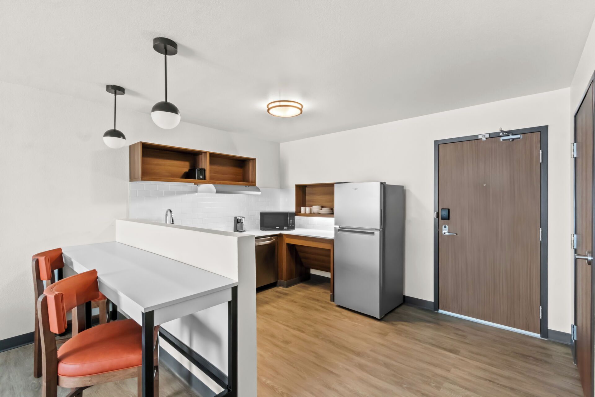 Een compacte keuken met een wit aanrecht, een ingebouwde magnetron, een roestvrijstalen koelkast en houten keukenkastjes. Er staat een eettafel met twee stoelen bij, met erboven een hanglamp.
